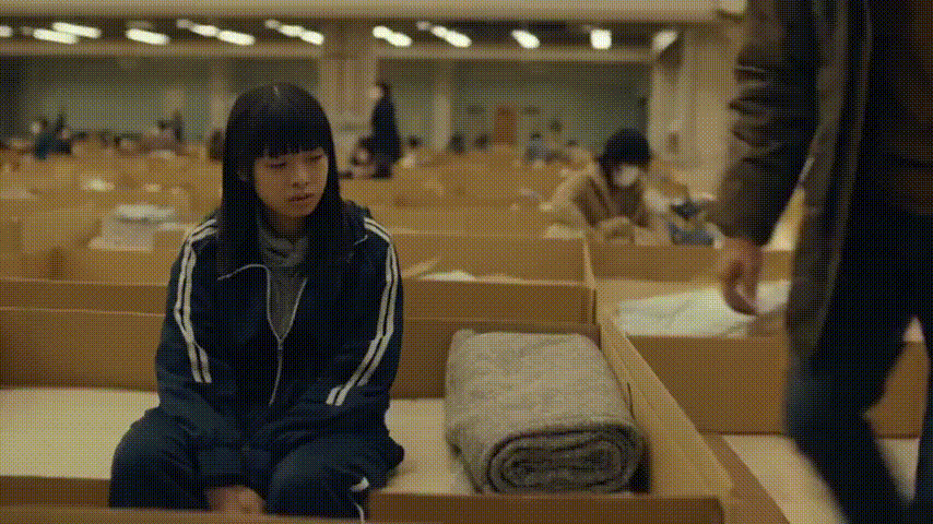 日本の被災地の避難所に多くの人が避難しています。ダンボールベッドがあります。そこに被災者の娘が座っています。表情は疲れています。ダンボールベッドだと分かるように少しカメラを遠ざけて下さい。娘の隣にお父さんが座ります。お父さんも疲れた表情です。二人とも体操着の上着です。お父さんが座ると二人は安堵した表情になります。